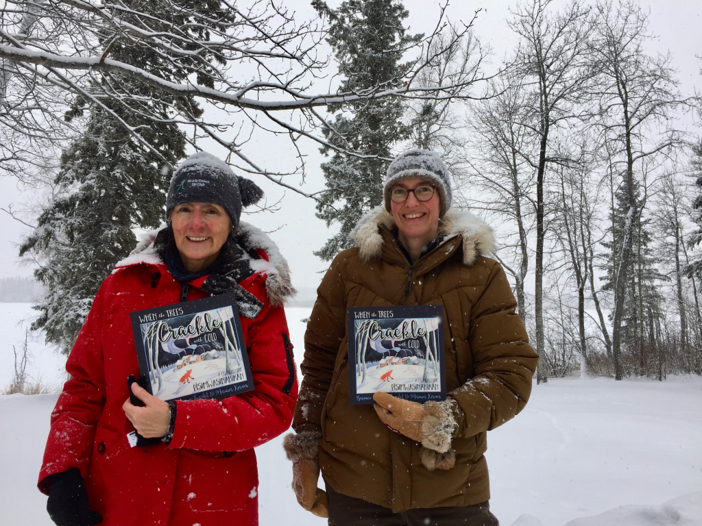 This is a photo of two people, outside in the snow with trees in the background. They are each holding a copy of their book "When the Trees Crackle with Cold: A Cree Calendar". On the left is Bernice Johnson-Laxdal, author of the book. On the right is Miriam Körner, illustrator and co-author of the book.