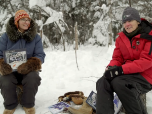 This image is a screenshot of the trailer video for the livestream reading of "When the Trees Crackle with Cold: A Cree Calendar" by Bernice Johnson-Laxdal and Miriam Körner. Miriam is on the left and Bernice is on the right in this picture. They are each sitting on a log, outdoors in the snow, with trees behind them.