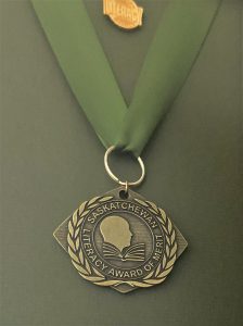 This is a photo of the medallion for the Saskatchewan Literacy Awards of Merit. The medallion has an image of a person and a book, with the words "Saskatchewan Literacy Award of Merit" around it in a circle. The bronze medallion is hung by a brass ring through an olive-coloured ribbon. There is a "Literacy" pin placed above it, all on a dark green framing matte.