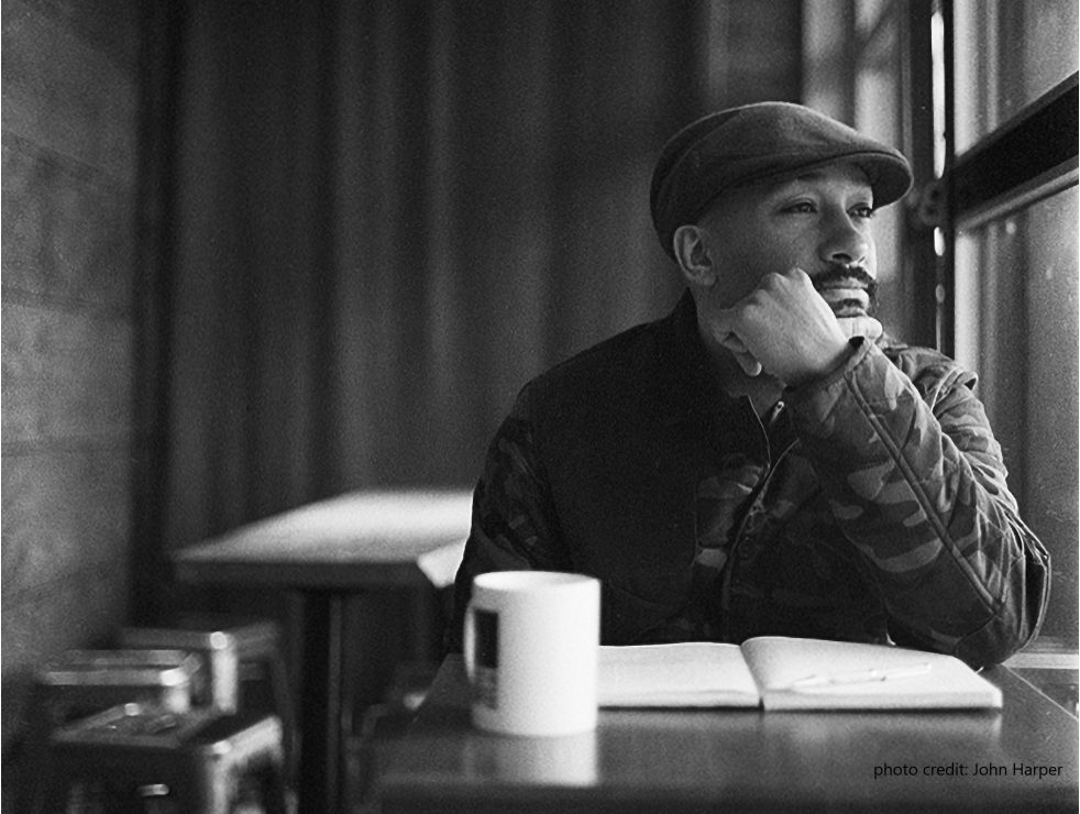 This is a photo of Khodi Dill by John Harper. Public speaker, writer, educator, and spoken word artist Khodi Dill sits at a table with a book and mug in front of him. He holds his hand up to his face and looks out the window to his left.