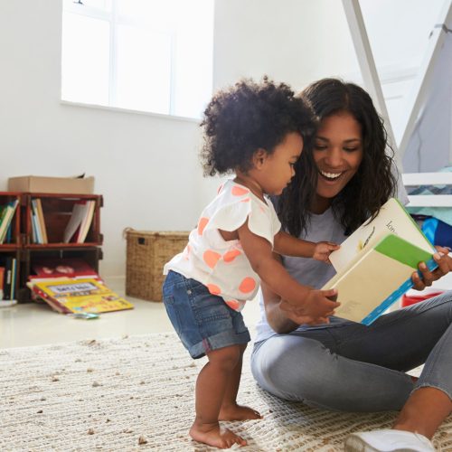 A mother and her baby daughter reading a book together in a playroom. They are seated comfortably on a soft rug, with the mother holding the book and the daughter looking at the pictures with curiosity.