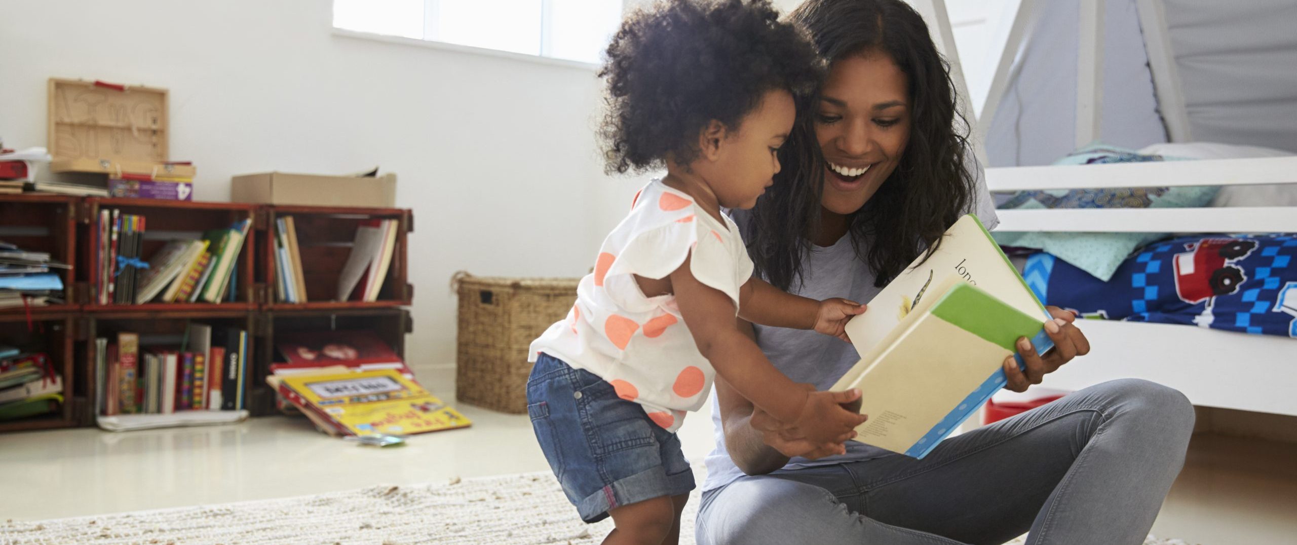 A parent and young child are reading a book together in a playroom. They are holding a book and reading together. The parent is smiling and the child is looking at the book with curiosity.