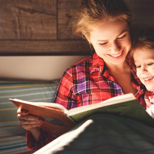 A mother and her child reading a book together in bed before going to sleep. They are under the covers, with the mother holding the book and the child snuggled close, looking at the pages with interest.