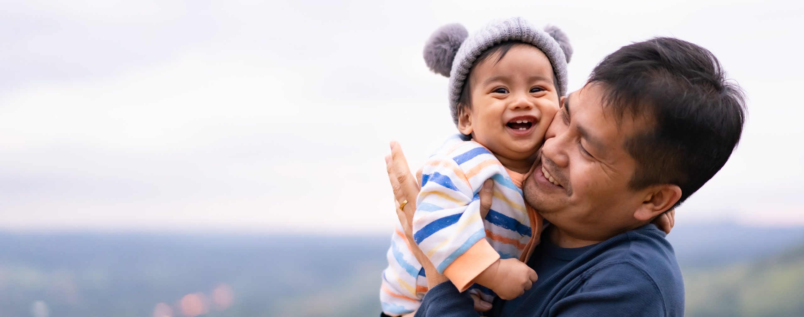 As we learn in the Rhymes, Songs, and Stories facilitator training, rhymes, songs, and stories happen everywhere in everyday life. This image shows an example of a parent holding their young child close as they share stories outside. Both the adult and the child are smiling from this time of bonding and learning.