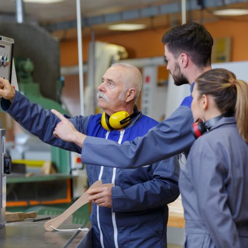 An older man and an adult working together with automated machinery in a workshop. The older man is providing guidance or instruction, while the adult is operating or adjusting the machinery. Both are focused on the equipment, with safety gear and technical tools in use.